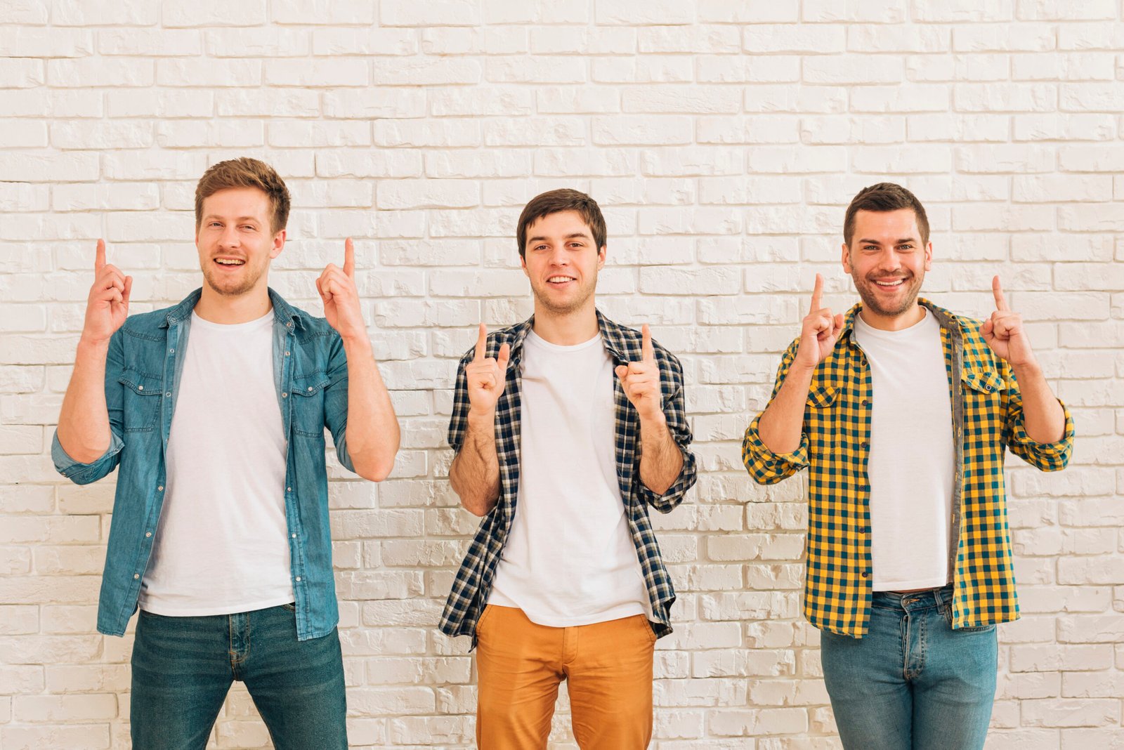 three-male-friends-standing-against-white-wall-pointing-finger-upward-looking-camera